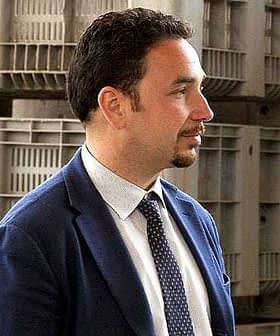 Profile view of a man wearing a suit and tie, standing in front of storage containers. - Olive Oil Times