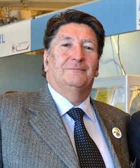 A man wearing a grey suit and a blue tie, standing in an indoor setting with a neutral expression. - Olive Oil Times