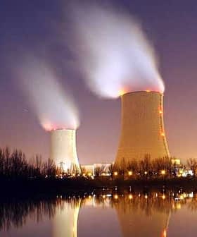 Two cooling towers emitting steam at night with reflections on the water surface. - Olive Oil Times