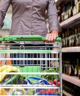 Person pushing a shopping cart filled with groceries in a supermarket aisle with shelves of bottles. - Olive Oil Times