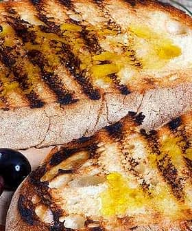 Two slices of grilled bread topped with olive oil and grill marks on a wooden board. - Olive Oil Times
