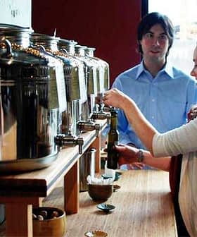 A woman dispensing oil from a large metal container at a service station while a man observes. - Olive Oil Times