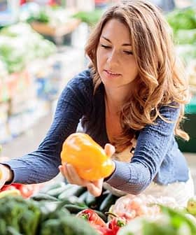 Woman holding two yellow bell peppers while shopping at a market filled with various vegetables. - Olive Oil Times