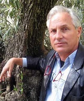 A man in a leather jacket leaning against the trunk of an olive tree with glasses in hand. - Olive Oil Times