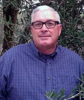 Man wearing glasses and a blue checkered shirt standing among olive trees. - Olive Oil Times