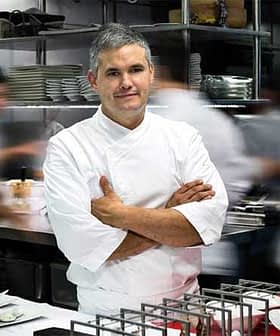 A chef standing with arms crossed in a busy professional kitchen environment. - Olive Oil Times