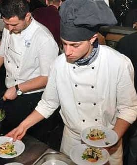 Two chefs in white uniforms plating food in a busy kitchen environment. - Olive Oil Times