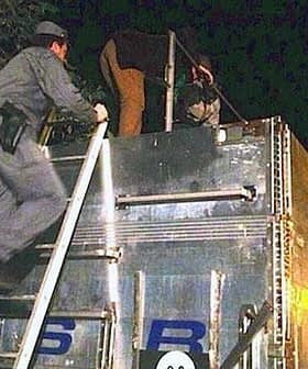 Two law enforcement officers climbing a ladder to inspect the top of a truck at night. - Olive Oil Times
