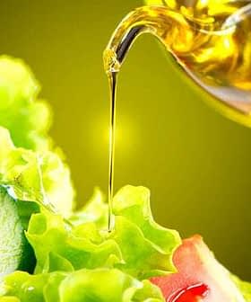 Olive oil being poured from a glass container onto a fresh green salad with lettuce and vegetables. - Olive Oil Times