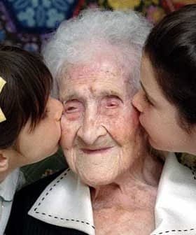 An elderly woman is being kissed on the cheeks by two young girls on either side of her. - Olive Oil Times