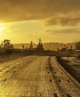 A wet road with reflective surfaces during sunset, featuring distant trees and signage. - Olive Oil Times