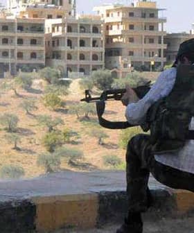 Individual in military attire aiming a firearm while crouched down in a rural area with buildings in the background. - Olive Oil Times