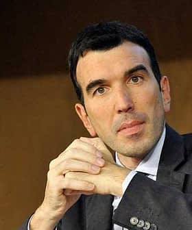 A man in a dark suit with short black hair, sitting with his hands clasped in front of him. - Olive Oil Times