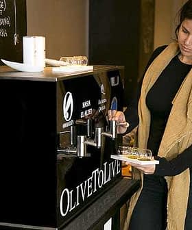 A woman dispensing olive oil from a black dispenser labeled 'OLIVETO LIVE' in a casual setting. - Olive Oil Times