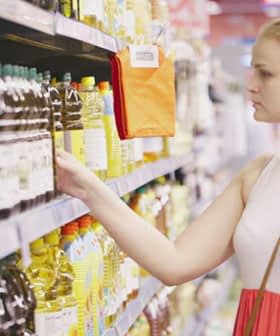 Woman with long hair in a white and pink dress reaching for a bottle of olive oil on a grocery store shelf. - Olive Oil Times