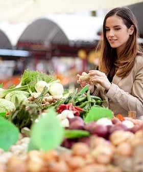 A woman examining fresh vegetables at a market stall filled with various produce. - Olive Oil Times