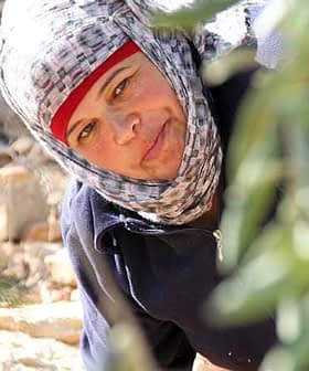 Woman wearing a headscarf and a dark jacket, smiling while harvesting olives in a field. - Olive Oil Times