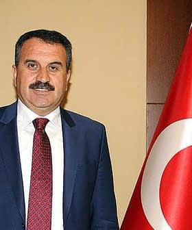 A man in a suit standing next to a Turkish flag, with a neutral expression and a mustache. - Olive Oil Times