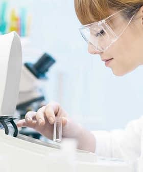 Female scientist in a lab coat and safety glasses operating laboratory equipment with test tubes. - Olive Oil Times