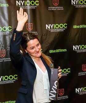 Woman with long hair raising her hand while holding an award at a culinary event backdrop. - Olive Oil Times