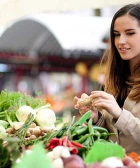 Woman examining fresh vegetables at a market stall with various produce visible. - Olive Oil Times