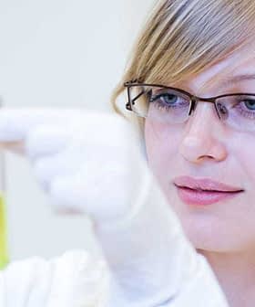 Woman wearing glasses and a lab coat holding a test tube with yellow liquid in a laboratory setting. - Olive Oil Times