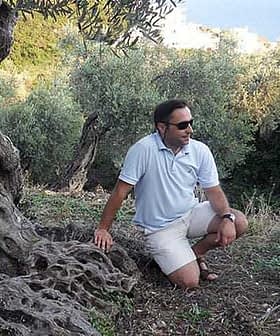 A man wearing sunglasses and a light shirt kneeling beside an olive tree in a natural setting. - Olive Oil Times