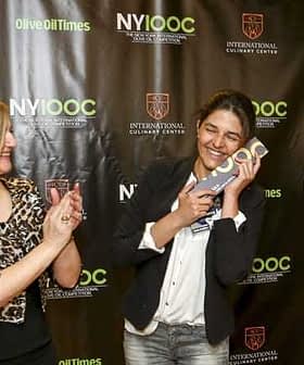 A woman smiling while holding an award at an event with a backdrop featuring Olive Oil Times and NYIOOC logos. - Olive Oil Times