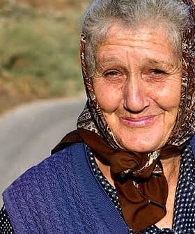 Elderly woman wearing a patterned headscarf and a blue vest, smiling at the camera. - Olive Oil Times