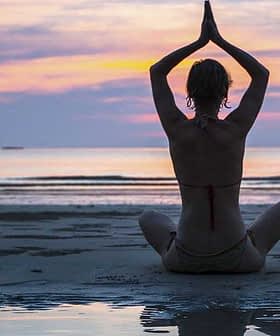 Individual sitting in a yoga pose on the beach during sunset with hands raised. - Olive Oil Times