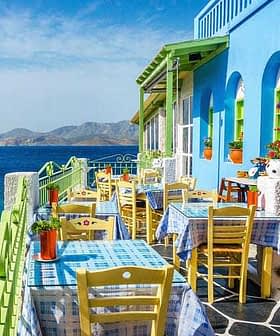 Outdoor dining area featuring tables and chairs in front of a blue and green building by the sea. - Olive Oil Times