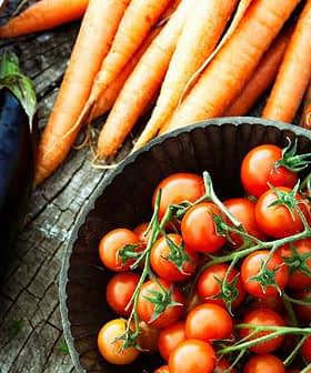 Bowl filled with cherry tomatoes surrounded by carrots and an eggplant on a wooden surface. - Olive Oil Times