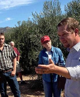 A group of people gathered in an olive grove, with one person speaking to the others. - Olive Oil Times