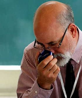 A man with glasses and a beard holding a blue object close to his face in a classroom setting. - Olive Oil Times