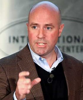 A man in a brown blazer and black sweater speaking at a podium during a conference. - Olive Oil Times