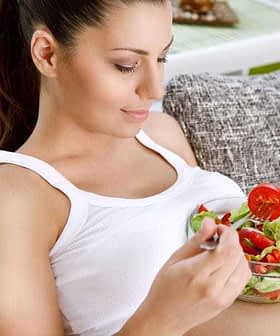 A woman sitting on a couch holding a bowl of salad with a fork, wearing a white tank top. - Olive Oil Times