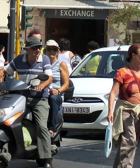 Two individuals on a scooter and a woman walking on a city street with vehicles in the background. - Olive Oil Times