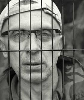 A man wearing glasses and a cap, looking through metal bars with a serious expression. - Olive Oil Times