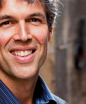 A man with gray hair and a suit smiling at the camera in an outdoor setting. - Olive Oil Times