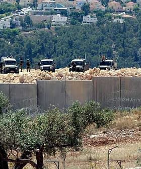 Two military vehicles stationed near a concrete barrier with barbed wire in a hilly area. - Olive Oil Times