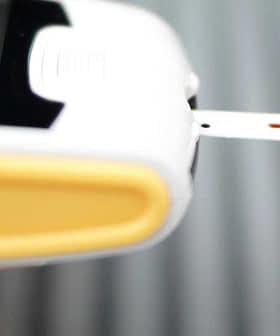 Person using a blood glucose meter with a test strip inserted and a drop of blood on their fingertip. - Olive Oil Times