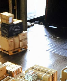A warehouse interior with stacked packages and a worker using a pallet jack to move boxes. - Olive Oil Times