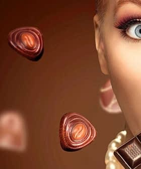 Woman with light skin holding a piece of chocolate with various chocolate shapes around her. - Olive Oil Times