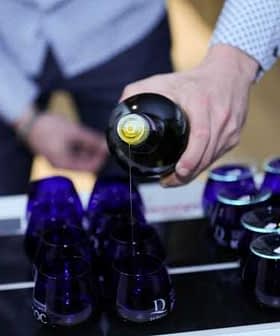 A person pouring olive oil from a bottle into a row of blue glass containers. - Olive Oil Times