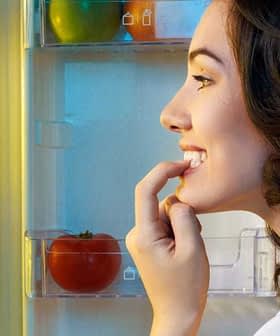A woman with dark hair smiling while looking into an open refrigerator filled with food items. - Olive Oil Times