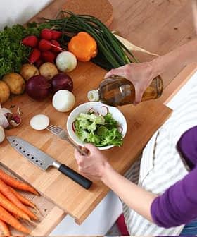 Individual pouring oil over a bowl of salad with various fresh vegetables on a wooden countertop. - Olive Oil Times