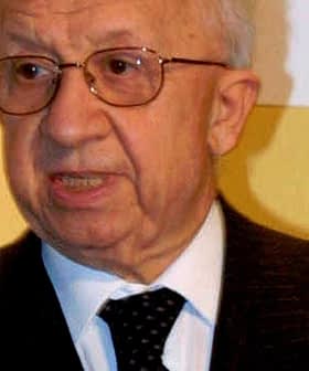 Elderly man wearing glasses and a dark suit speaking during an event. - Olive Oil Times