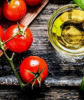 Fresh red tomatoes, green basil leaves, and a small glass of olive oil on a wooden surface. - Olive Oil Times