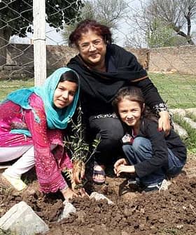 A woman and two children planting a small tree in a garden area with a fence in the background. - Olive Oil Times