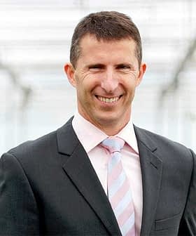 Man wearing a dark suit and light blue striped tie, smiling in a greenhouse setting. - Olive Oil Times